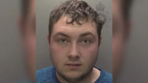 Humberside Police A police custody photo of a man - Dylan Lowther - looking at the camera. He has short brown hair and short brown facial hair. He is wearing a blue top. The wall behind him is grey.