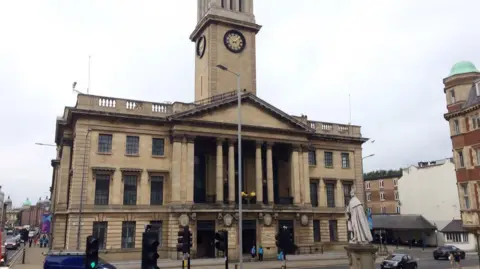 A large historic building with classical architecture, featuring tall entrance columns and a central clock tower. The stone structure has multiple windows. In front of the building stands a statue on a pedestal, with traffic lights and vehicles visible on the surrounding street.