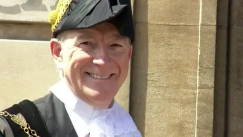 BBC A man wearing ceremonial clothing featuring a black gown with gold chains and a white lace collar. The background features the stone wall of Hull's Guildhall.