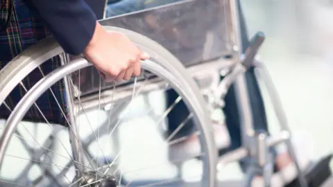 Getty Images A silver wheel of a wheelchair can be seen with a hand on it about to move it forward. You can see in blurred vision the person's legs and feet sat on the rests at the front of the wheelchair.
