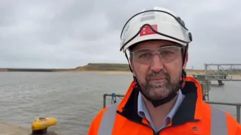 Andrew Turner/BBC Richard Goffin, wearing an orange high visibility jacket, white hard hat, with safety visor and safety glasses. Beside him are port equipment, and beyond, the spending beach and unused area of the Great Yarmouth outer harbour.