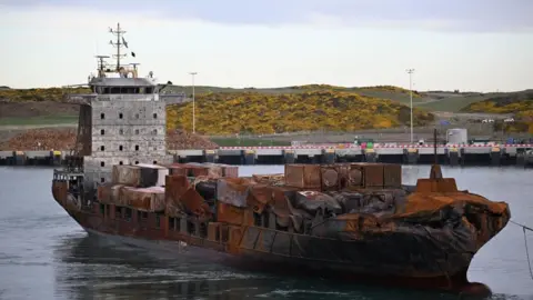 PA Media A container ship with most of its hull and its containers damaged by fire. In the background is a quay.