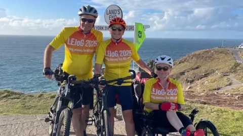 NHS Humber Health Partnership Three people in front of a sign marked "Land's End". A man and a woman are standing and holding bikes. A woman on the right is sitting on a trike. They are all wearing yellow and orange cycling kit with white writing on it, cycling helmets and sunglasses. The sea is in the background. The sky is blue and cloudy.