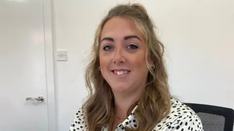 A woman with long curly blond hair smiles at the camera as she sits in a black chair. She is wearing a white and black polka-dot shirt.