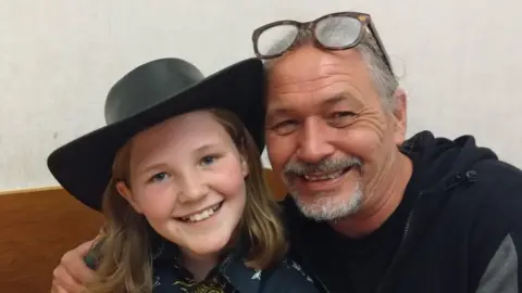 Supplied A man hugs his son as they sit on a sofa and smile towards the camera. The man has grey hair, tied back in a pony tail, and a neatly cropped moustache and beard. His glasses sit on his forehead. The son has shoulder-length dark-blonde hair and is wearing a black cowboy hat.