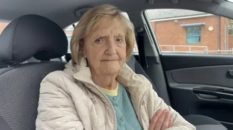 BBC A close up shot of an elderly woman wearing a beige coat with a blue jumper underneath. She has her arms crossed. She has short blond hair. She is sat in her car.