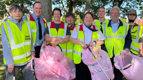 BBC A group of eight men and women wearing high-visibility vests and carrying pink litter bags and long litter-picking grabbers. They are standing in a park in front of trees.