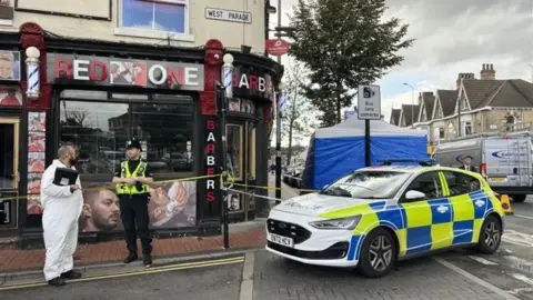 BBC Police at the scene of the reported incident. The picture shows a police car in front of a blue police tent. There are two police officers in the picture - one wearing a police uniform and the other wearing a white hazmat suit. An area outside a shop called Red One Barbers is cordoned off with blue and white police tape. 