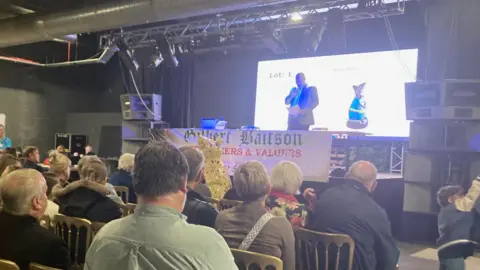 BBC The auctioneer stands in front of a screen that displays one of the rat sculptures. People are sat on chairs with their backs to the camera during the bidding.