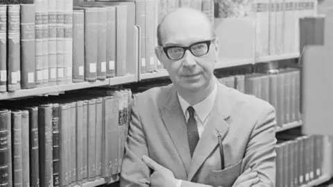Getty Images A black and white archive shot of a man standing in front of shelves full of books. He is wearing black-rimmed spectacles, as well as a shirt, a tie and a suit.