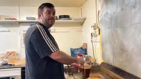 BBC Craig Beverley, the owner of Bankside Cafe, is stood in a kitchen and stirs a jug filled with hot liquid as he looks at the camera. Mr Beverley has dark brown hair, a cropped beard and moustache. He is wearing a navy blue T-shirt with three white track lines on the sleeve.