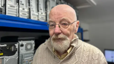 A man with white eyebrows and a white mid length beard stood next to a series of refurbished computers. He has a cream V neck sweater on with a yellow shirt underneath, he has small circular glasses on and is looking at the camera about to explain how the computers operate
