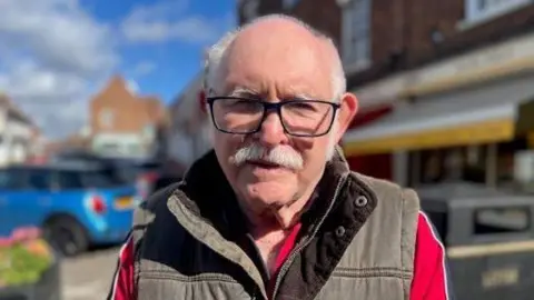 BBC A head and shoulders shot of a man - Keith Pollard - stood in a street looking at the camera. He is wearing a red top, a brown bodywarmer and glasses. He has short grey hair and a moustache.