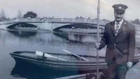 BBC/Unknown A black and white photo showing a man wearing a dark uniform and peaked cap. He is holding a large boating pole and standing in front of rowing boats at a large lake in a park. In the background a double-arched, stone footbridge crosses the lake.
