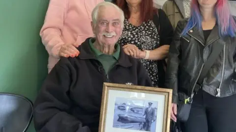 BBC Stuart MacDonald smiling at the camera wearing a black work fleece and a green shirt, and holding a frame with an old black-and-white photo of him. A group of women, partially out of frame, stand behind him, with one resting a hand on his shoulder.