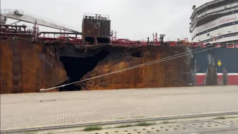 Andrew Turner/BBC The significant hole in the side of the Stena Immaculate, at rest on the North terminal of Great Yarmouth's outer harbour. The hull is rusting and fire damaged following a collision in March off the Humber Estuary. The ship superstructure and bridge is visible to the right of the image. Quayside hardstanding is visible in the foreground.