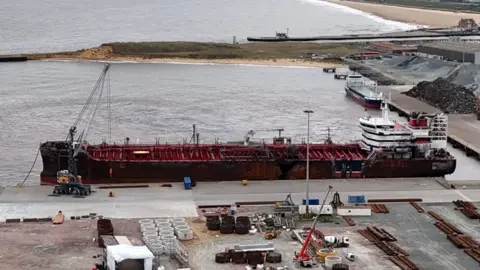 Qays Najm/BBC A drone image looks down on the red supertanker at anchor in a port, with equipment, machinery and materials on the land around it.