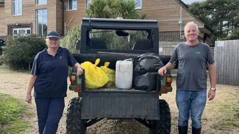 Norfolk Wildlife Trust A woman in dark clothing and a cap stands to the left of a beach buggy parked on sandy ground, with the tailgate of the vehicle open and showing bagged waste which has been gathered, and man in a grey t-shirt and jeans stands to the right with a wooden cladded building in the background.