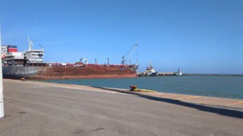 Supplied The Stena Immaculate rests on the North terminal of Great Yarmouth's outer harbour, with a tug at the bow. The hull is rusting and fire damaged following a collision in March off the Humber Estuary. Another tug is further towards the right of the image with quayside hardstanding in the foreground.