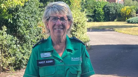 St John Ambulance Woman with short hair and glasses wearing a green St John Ambulance uniform. She is stood outside in front of a bush.
