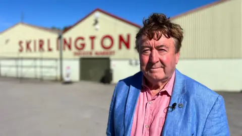 BBC A man standing in front of white sheds with "Skirlington" written in big red lettering on them. He has short dark hair and is wearing a pink shirt and a blue jacket.