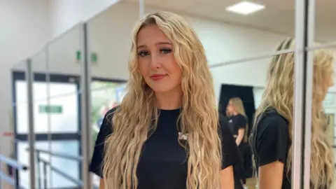 BBC Scarlett is stood next to a mirror, her reflection can be seen, as she smiles at the camera. The 17-year-old has long blonde wavy hair and is wearing a black T-shirt.
