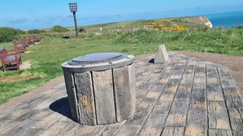 View of the toposcope. A stone circular platform with an inscribed metal plate on the top it is surrounded by rectangular stone flags. It sits on the top of Bempton Cliffs surrounded by grass and wooden benches with the sea in the background.