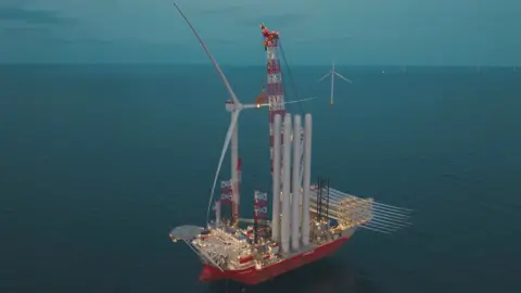 RWE Offshore Wind A ship laden with wind turbine components is at sea. An assembled wind turbine can be seen in the background.