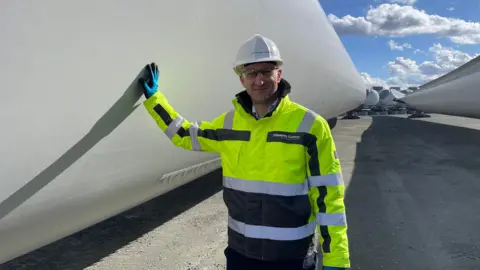 BBC A man wearing a high-visibility jacket and a protective helmet places his hand on the side of a large white wind turbine blade. 