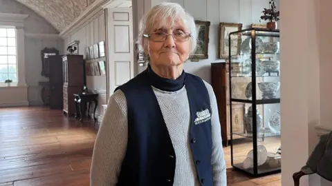 Head guide of Burton Agnes Hall, Pauline Waslin looks into the camera. She has short, white hair and silver-rimmed glasses. She is wearing a grey jumper with a navy-blue unbuttoned unic over top, it is branded with 'Burton Agnes Hall' with a small white drawing of the stately home.