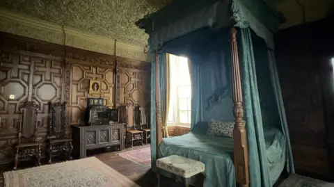 The Queen's state bedroom at Burton Agnes Hall. There is a large, wooden four-poster bed in the centre of the room, it has blue curtains and bedding. The walls are wood panelled in a geometric pattern. To the left of the bed, there are four wooden, high-backed chairs and a wooden dresser. There is a gold-framed painting on the wall of a boy in a red shirt.