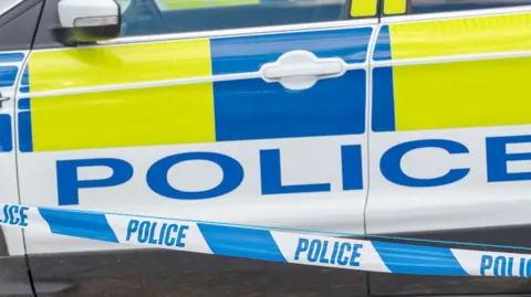 BBC Close up of the side of a police car with tape in front of it.
