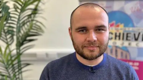 BBC A man wearing a blue jumper stares into the camera. They have short dark hair and a beard. A green plant is visible in the background as well as a backdrop which illustrates the words 'achieve' describing some of the aspirations at the Warren Youth Project.