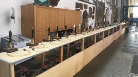 Hull and East Yorkshire CAMRA A long line of more than 20 beer pumps attached to a long wooden temporary bar inside a church. Old stonework and windows can be see in the background. 