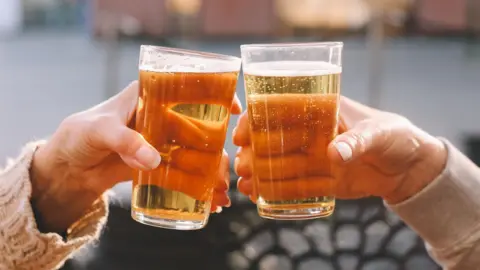 Getty Images A close-up of the hands of two people clinking pin glasses of amber-coloured beer together in front of an out-of-focus background.