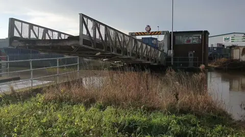 East Riding of Yorkshire Council A metal lifting bridge in operation. The single-track bridge has grey railings and is half raised over a brown river, with reeds and foliage in the foreground. In the background there is a square, brick-built control room and a height barrier with black and yellow hazard sign. 