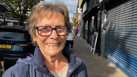 A woman standing on a pavement smiling at the camera. She is wearing a blue jacket and glasses. There are shops with the shutters down in the background.