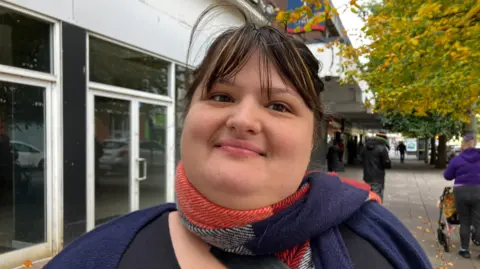 A woman standing on a pavement smiling at the camera. She has dark hair and a red and blue scarf around her neck. There are other people in the background and a glass-fronted building.