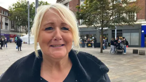 BBC A woman with blonde hair parted down the middle, wearing a black coat is standing in Hull city centre. There are shops behind her and an open square.