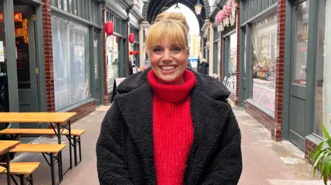 BBC Steph Ellis pictured standing inside the arcade, She has light-coloured hair and is wearing a black jacket and red jumper. She is smiling at the camera. There are shops on either side in the background.