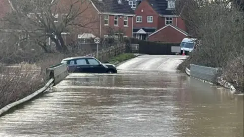 BBC A black mini part-submerged in floodwater on a road over a bridge.