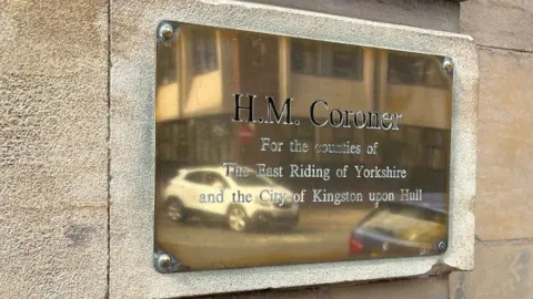 A brass plaque on the wall outside Hull Coroner's Court, with cars reflected in it. It reads "HM Coroner for the counties of the East Riding of Yorkshire and the City of Kingston upon Hull".