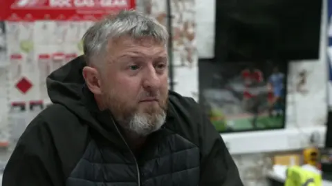 A man with grey short hair and a grey and brown short beard and moustache is looking to the side with a serious expression. He is wearing a black jacket and you can see a workshop blurred behind him.