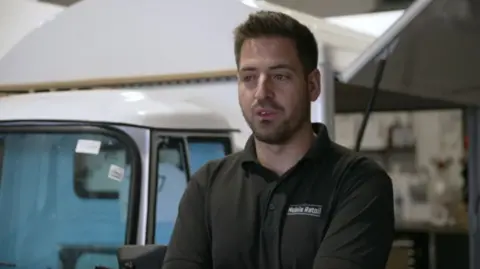 A man with short brown hair styled as a quiff is wearing a black polo shirt that says 'Mobile Retail on it'. Behind him you can see a white mobile van. He is looking to the side and has short brown stubble and a moustache.