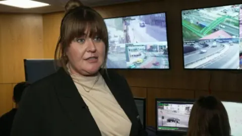 BBC A woman with a full brown fringe with her hair up in a bun. She is wearing a black blazer and white top and behind her are screens analysing traffic in a city.
