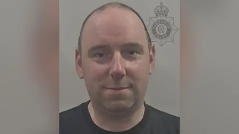 Humberside Police A man with very short hair which is bald at the front is smiling for a police photo. He is wearing a black t-shirt and the background is grey.