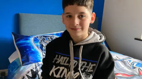Teenage boy with dark hair wearing a Sheffield Steelkings hoody and sitting on his bed which has blue, white and red Sheffield Steelkings bed covers
