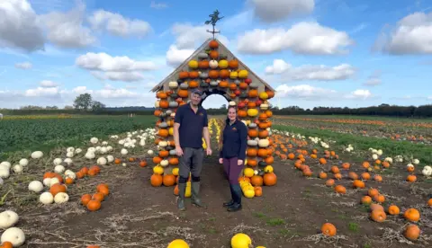 BBC / Isabella Norrison Man in blue t-shirt and blue cap and dark trousers. He is wearing wellington boots and smiling. He is stood with a woman with sunglasses on her head, blue fleece, grey top and maroon trousers. She is wearing wellington boots and smiling. There are lots of pumpkins that are white, yellow and orange in the background. There is a house shaped structure with pumpkins on it and a weather vane on top.