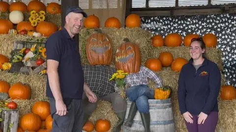 BBC / Isabella Norrison Man in blue t-shirt and blue cap and dark trousers. He is smiling. He is stood with a woman with sunglasses on her head, blue fleece, grey top and maroon trousers. She is smiling. There are lots of pumpkins in the background.