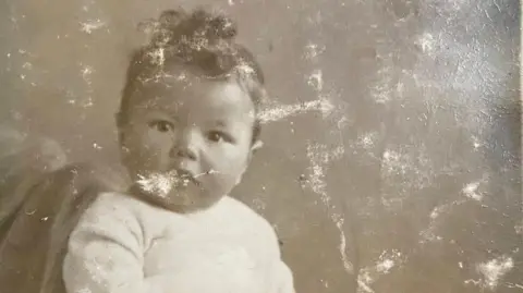 Kizzie Elliott An old sepia photograph of a young child. The boy looks directly at the camera and has dark curly hair and large brown eyes. He wears a white jumper and the picture is cracked white and appears weathered. 